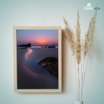 A serene beach sunset with vibrant colors reflecting off the water, framed by rocks along the shoreline.